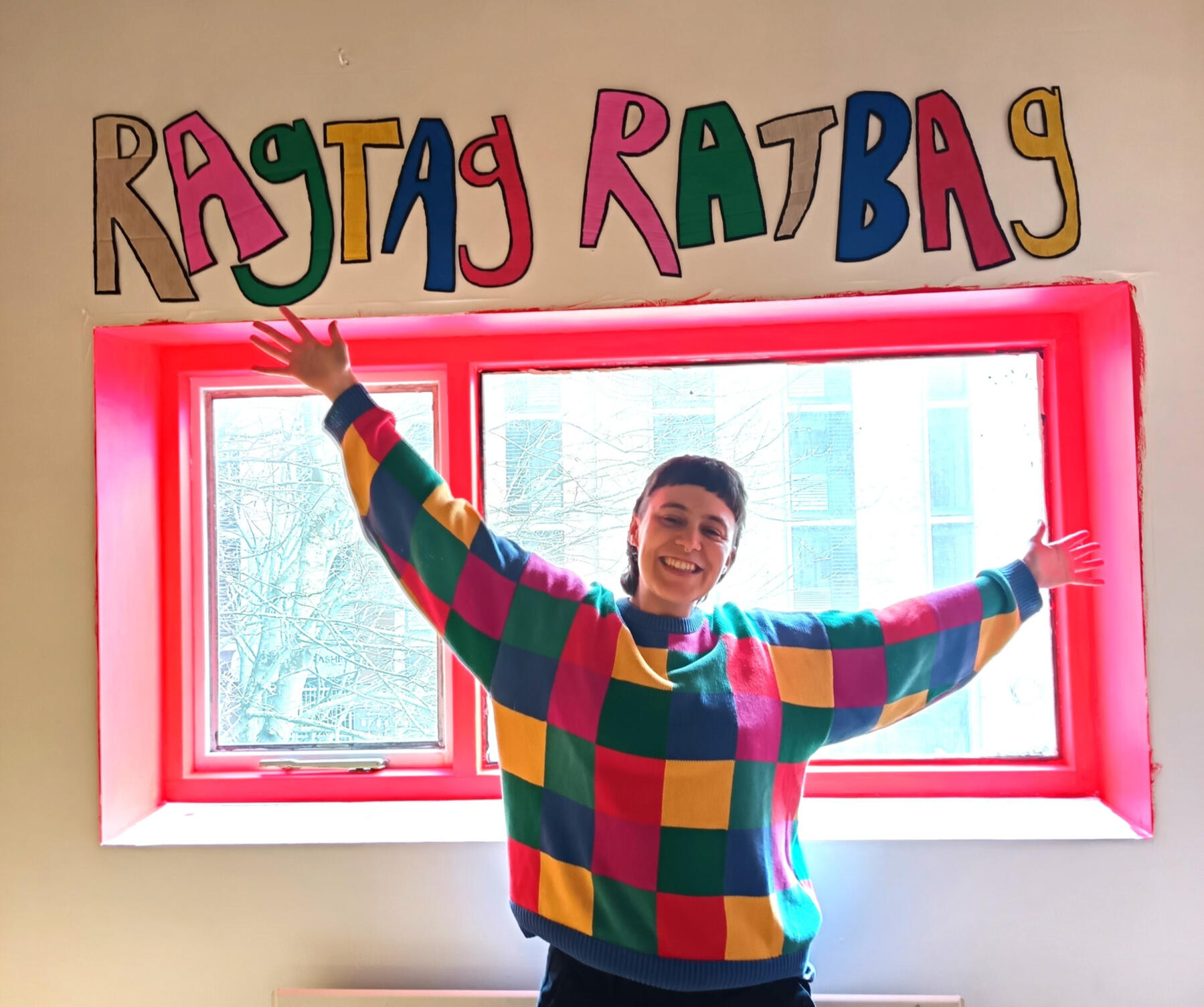 photo of Evie, who is wearing a colourful patchwork chequered sweater, has short hair and is stood infront of a red window frame with their arms in the air, smiling, above them on the white wall it reads "Ragtag Ratbag" in big colourful letters with a blac